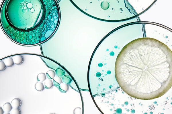 Abstract close-up of overlapping clear glass circular dishes containing turquoise and aqua liquid, bubbles, and a sliced white citrus wedge on a pale background.