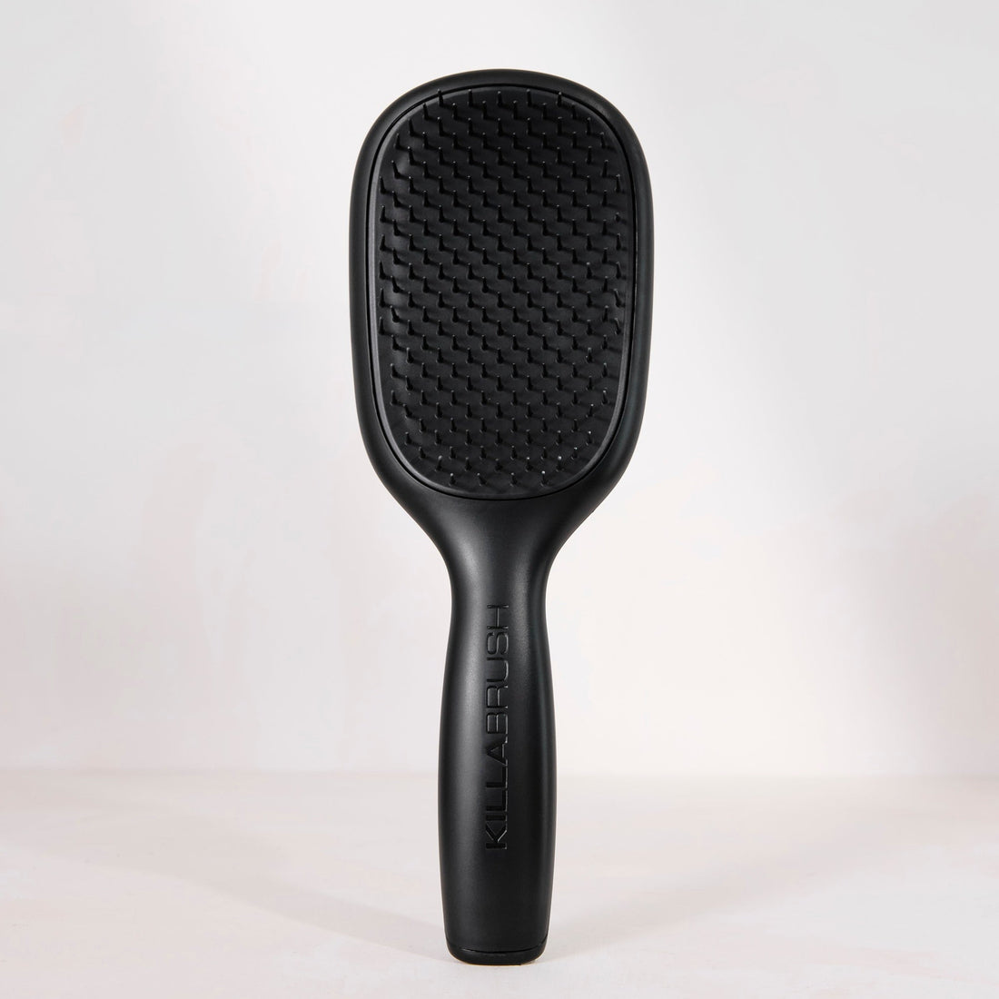 A black hairbrush with a textured paddle head and a slim handle, standing upright against a light gray studio background.