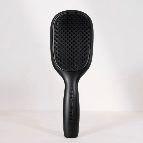 A black hairbrush with a textured paddle head and a slim handle, standing upright against a light gray studio background.