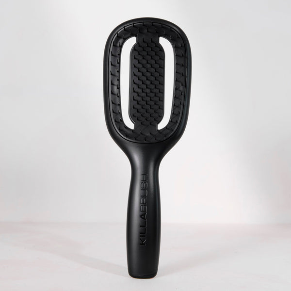 A black hairbrush with a textured, honeycomb-like bristle head and a sleek handle, standing upright against a light gray studio background. The brand name 'KILLABELUSH' is vertically embossed on the handle.