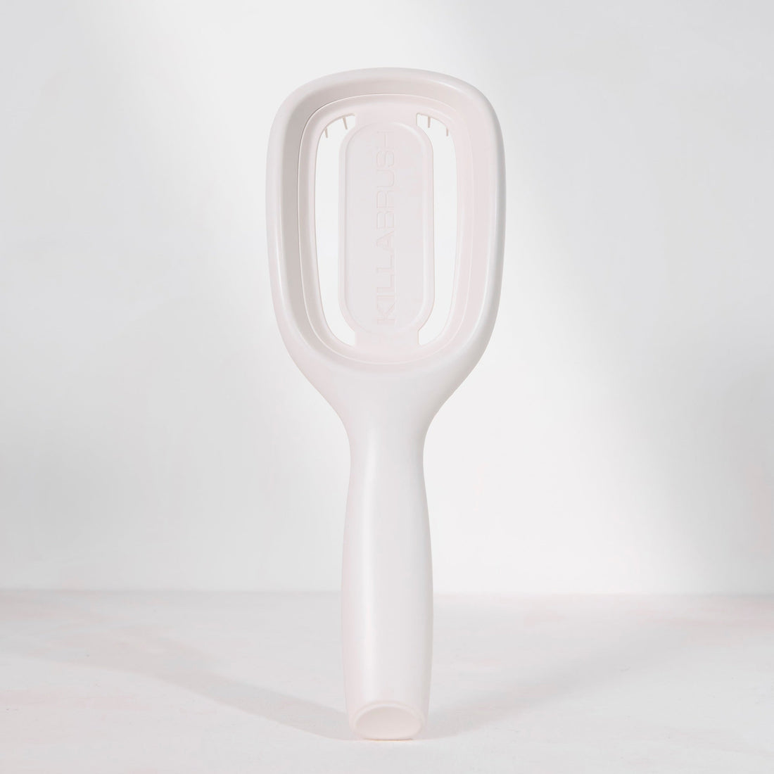A minimalist, matte white hairbrush with a rounded rectangular head and a smooth handle, standing upright on a light gray surface against a matching light gray background.
