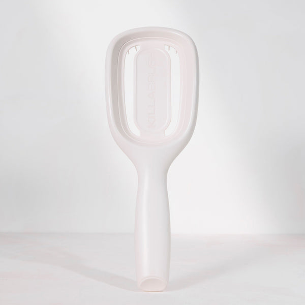 A minimalist, matte white hairbrush with a rounded rectangular head and a smooth handle, standing upright on a light gray surface against a matching light gray background.