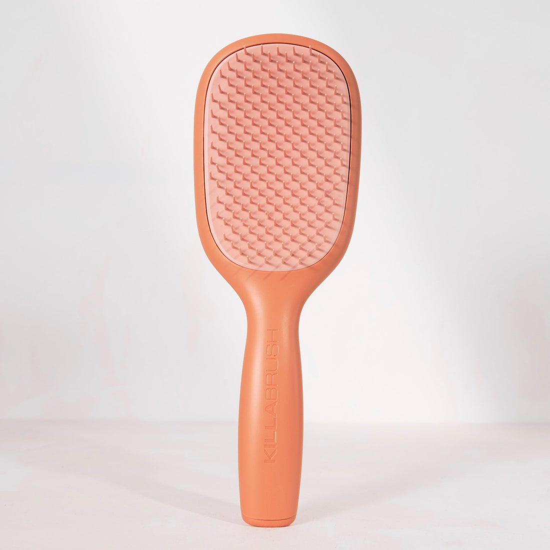 A single paddle-style hairbrush standing upright against a clean white studio background. The brush has a matte peachy-brown handle and frame with a lighter pink textured bristle pad.