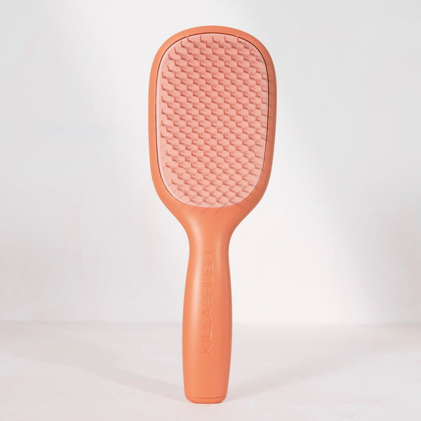 A single paddle-style hairbrush standing upright against a clean white studio background. The brush has a matte peachy-brown handle and frame with a lighter pink textured bristle pad.