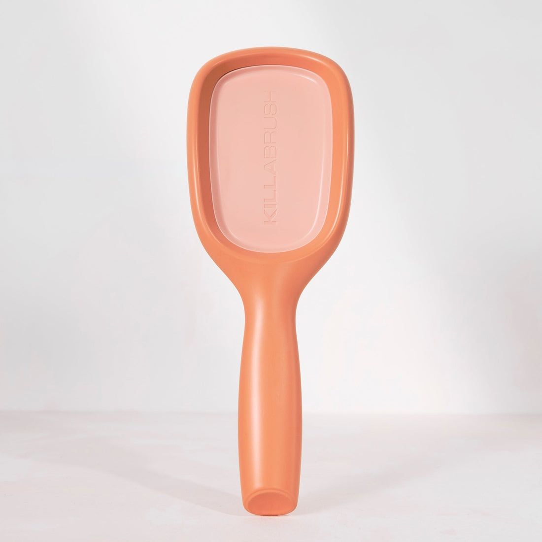 A single paddle hairbrush standing upright against a clean white studio background. The brush has a matte peachy-pink color with a slightly lighter pink face and subtle embossed branding.