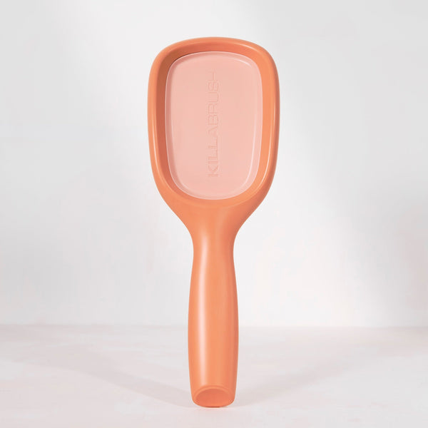 A single paddle hairbrush standing upright against a clean white studio background. The brush has a matte peachy-pink color with a slightly lighter pink face and subtle embossed branding.