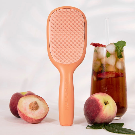 A peach-colored handheld grooming brush with a textured bristle head stands upright on a white surface, styled with whole and sliced peaches and a glass of iced tea with mint leaves.